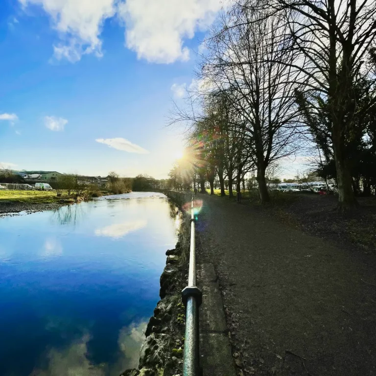 « Chemin de promenade bordé d’arbres le long d’une rivière qui reflète le ciel bleu, sous un soleil d’hiver. »